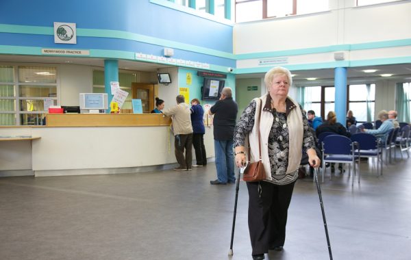 An older woman walking with sticks in a GP waiting area