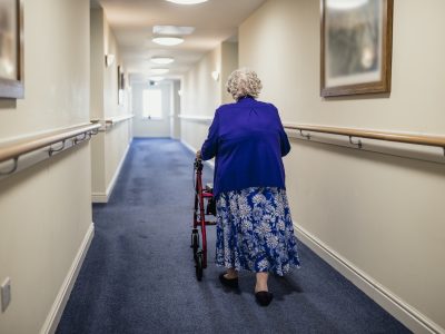 An elderly woman with a walking aid walks down a corridor in a care home