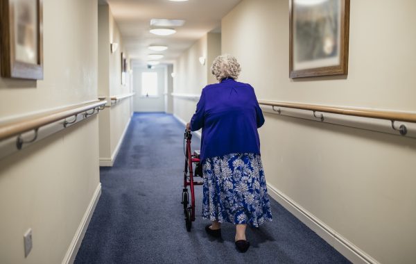 An elderly woman with a walking aid walks down a corridor in a care home