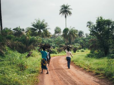 Three people walking along a dirt road flanked by palm trees and vegetation somewhere in Africa