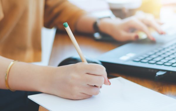 A woman is taking notes while working on her laptop
