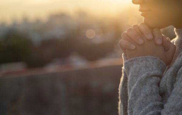 A woman clasps her hands in hope with the sunrise behind her