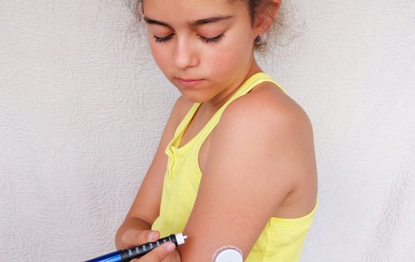 Girl stands and injects insulin from insulin pen in arm. On the arm is also placed white sensor for continuous glucose monitoring in blood