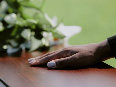 A black woman's hand rests on a coffin during a funeral, with flowers in the background
