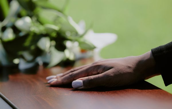 A black woman's hand rests on a coffin during a funeral, with flowers in the background